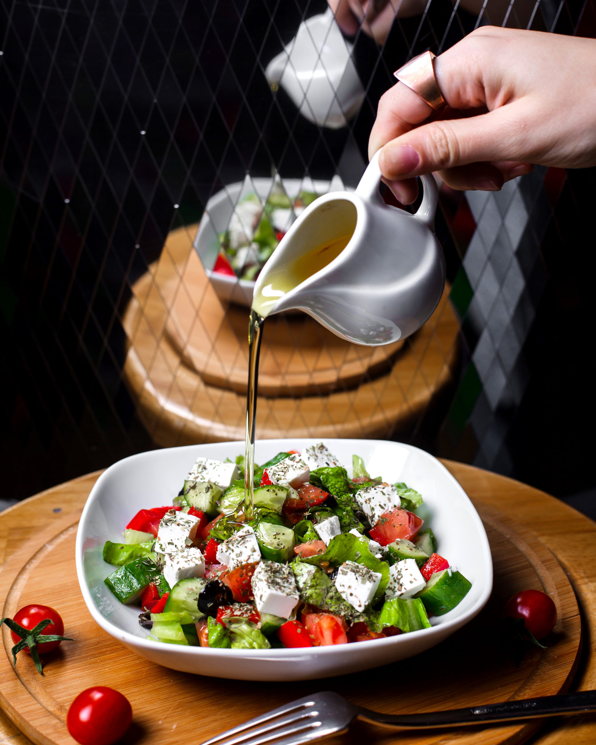 side view of hand pouring olive oil on fresh salad with feta cheese tomatoe cucumbers in a white bowl